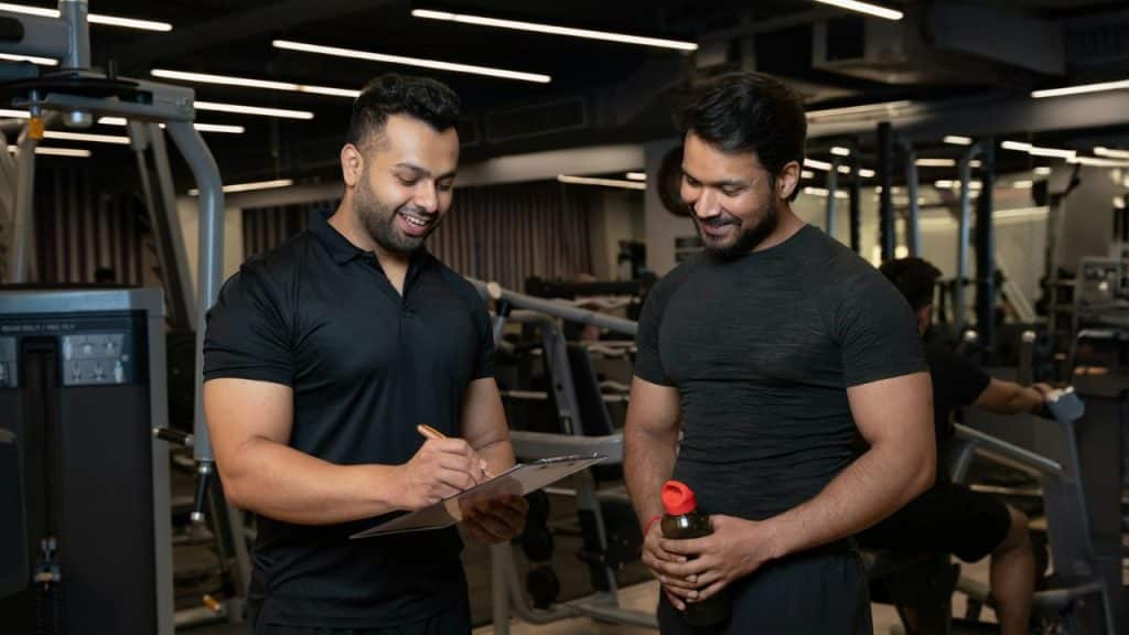 A personal trainer with a clipboard and a smiling client with a water bottle stand in a gym.