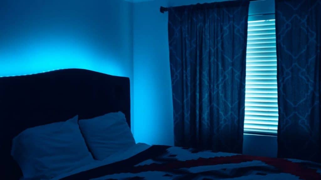 A dimly lit bedroom features a bed with blue accent lighting and dark curtains over a window.