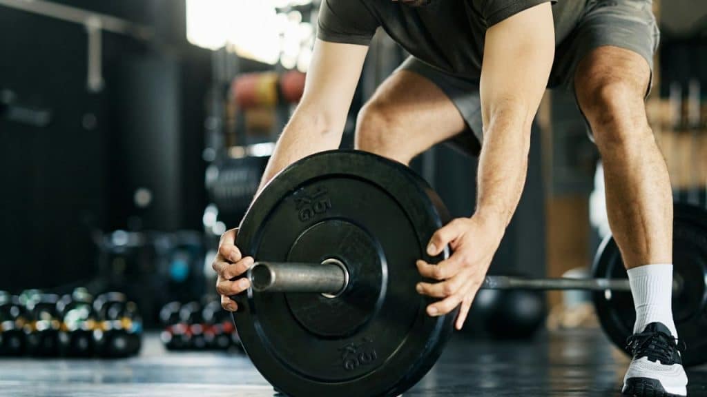 A person in a t-shirt and shorts places a black weight plate onto a barbell on a gym floor.