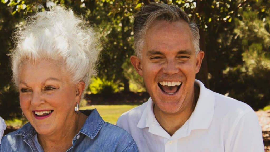 A man and woman with light-colored hair laugh outdoors under a tree.