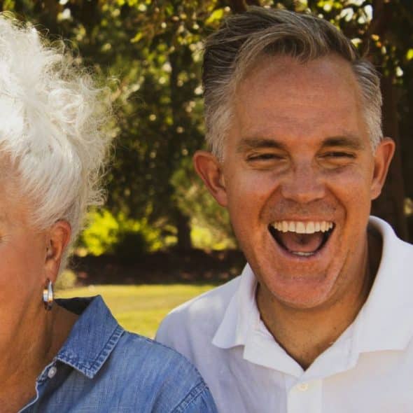 A man and woman with light-colored hair laugh outdoors under a tree.
