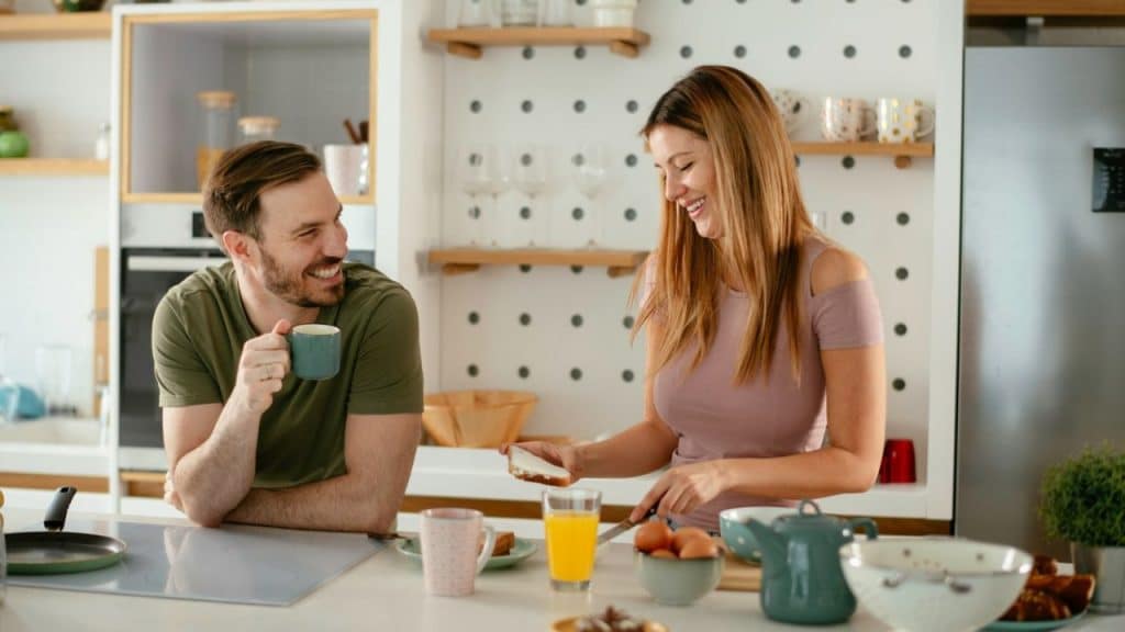 A man with a beard holds a mug and smiles at a woman who is preparing food in a kitchen.