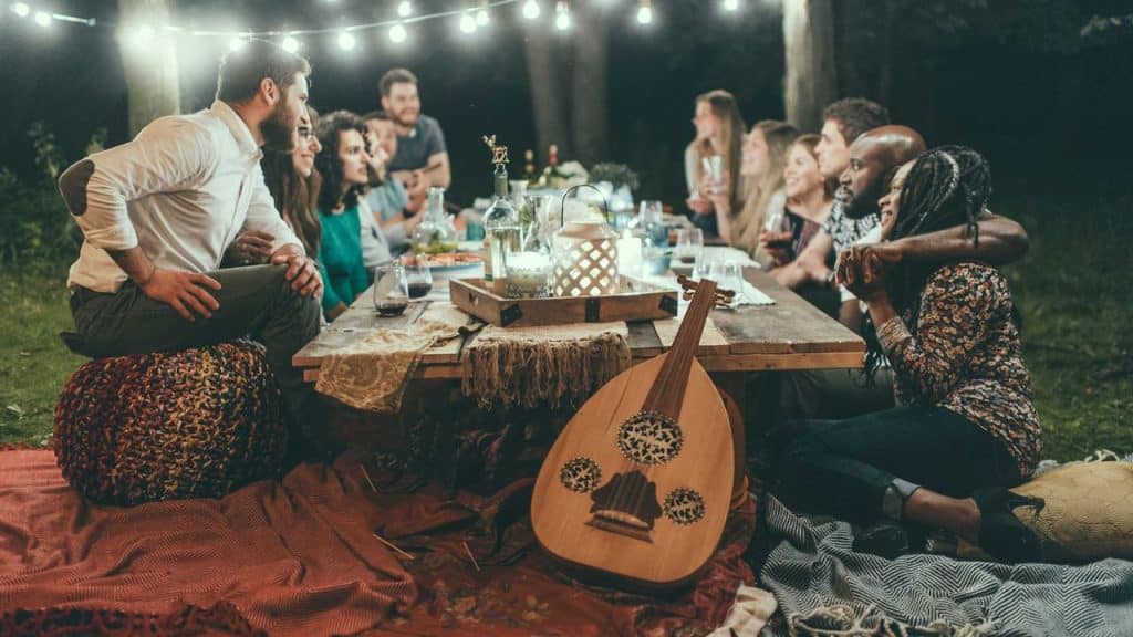 A group of friends enjoys an outdoor dinner party under string lights with a lute on the table.