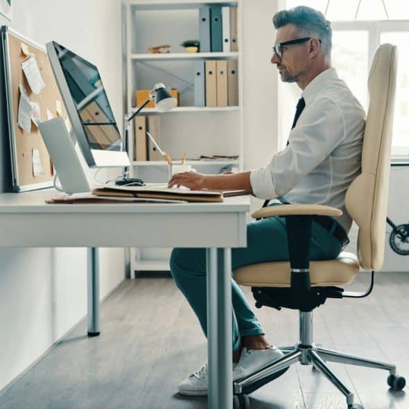 A man with glasses and gray hair sits at a desk, working on a computer.