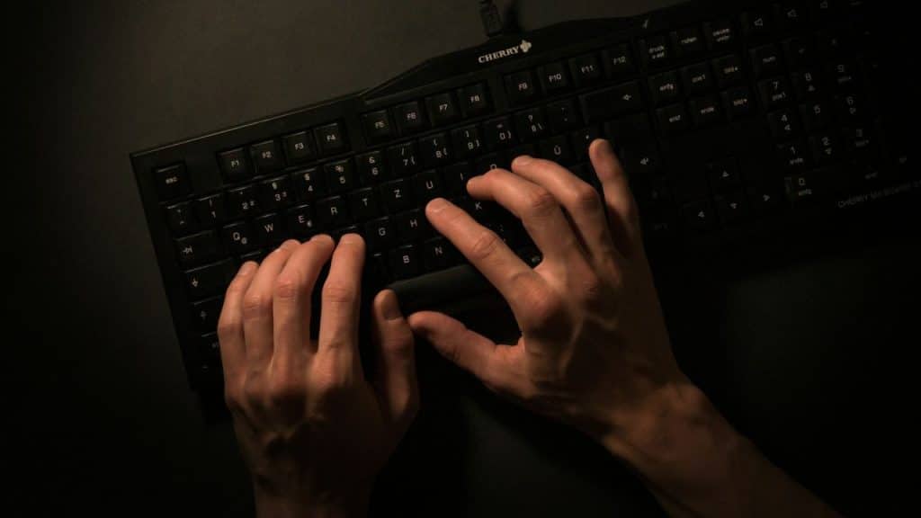 A person's hands are typing on a black computer keyboard in a dimly lit setting.