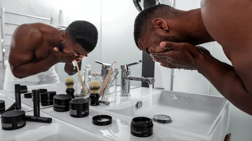 A shirtless man is washing his face at a bathroom sink surrounded by various grooming products.