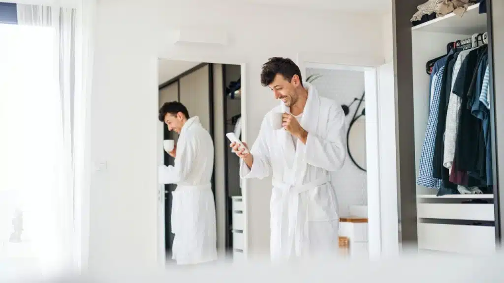 A smiling man in a white robe holds a phone and a mug, with his reflection visible in a mirror.