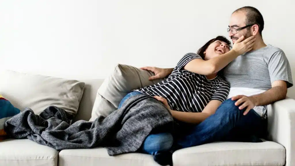 A man with glasses sits on a couch while a woman with a striped shirt lies on him, both smiling.