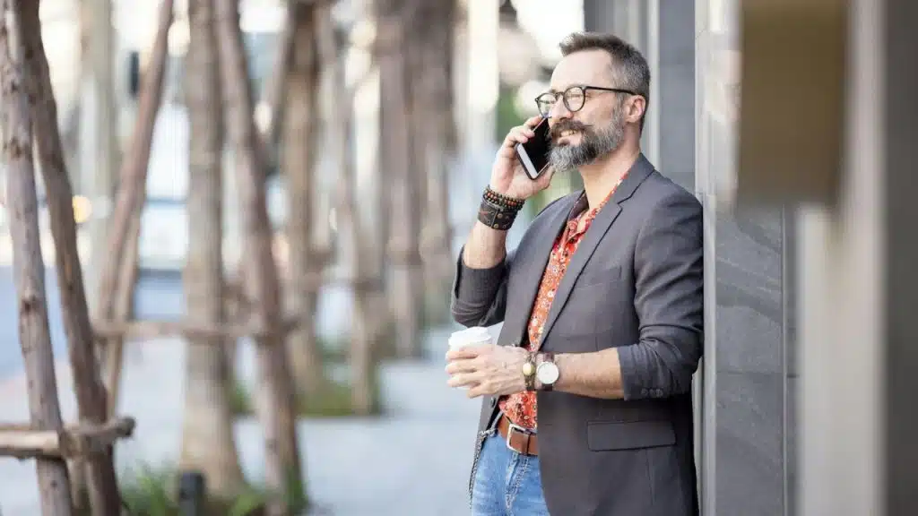 A stylish man with a beard and glasses smiles while talking on a phone and holding a coffee cup outdoors.