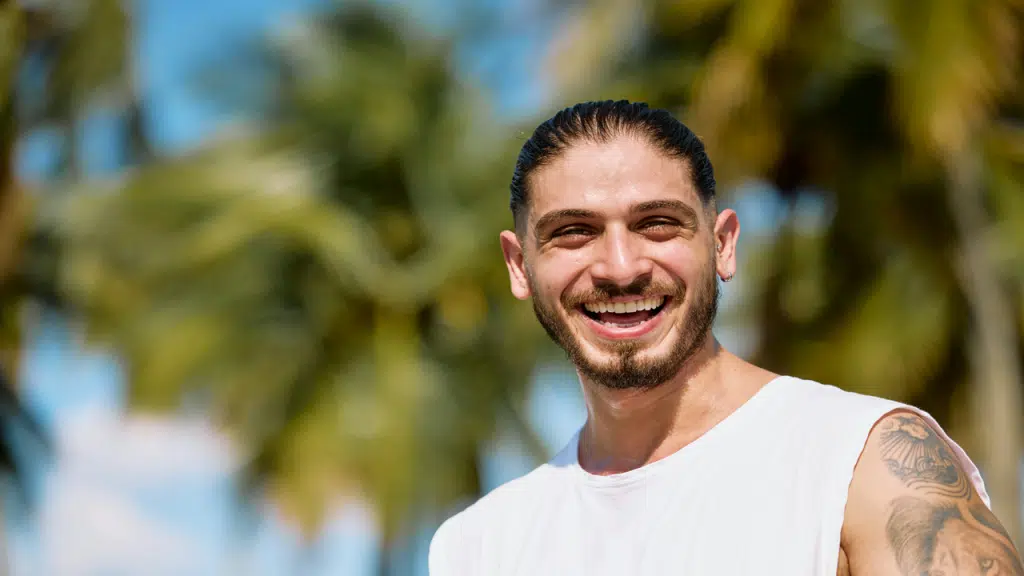 A smiling man in a sleeveless white shirt stands outdoors on a sunny day with palm trees blurred in the background.