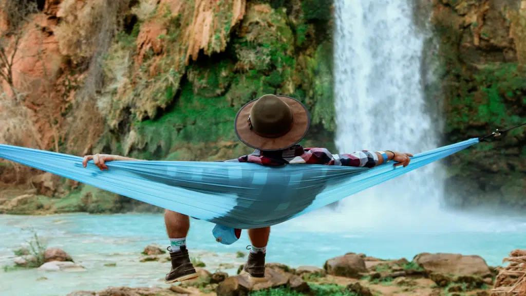 A person wearing a hat and a flannel shirt relaxes in a blue hammock suspended near a turquoise pool with a waterfall in front.