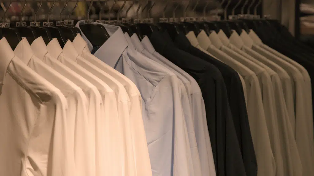 A row of neatly hung dress shirts in shades of white, light blue, and black is displayed on hangers.