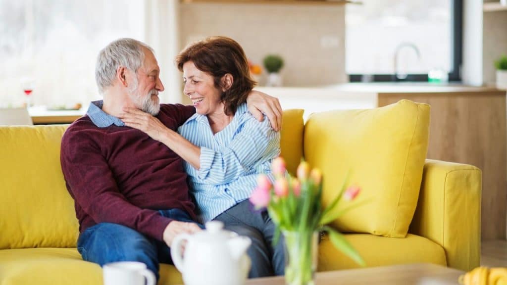 An older couple smiles and hugs on a yellow couch in a brightly lit room.