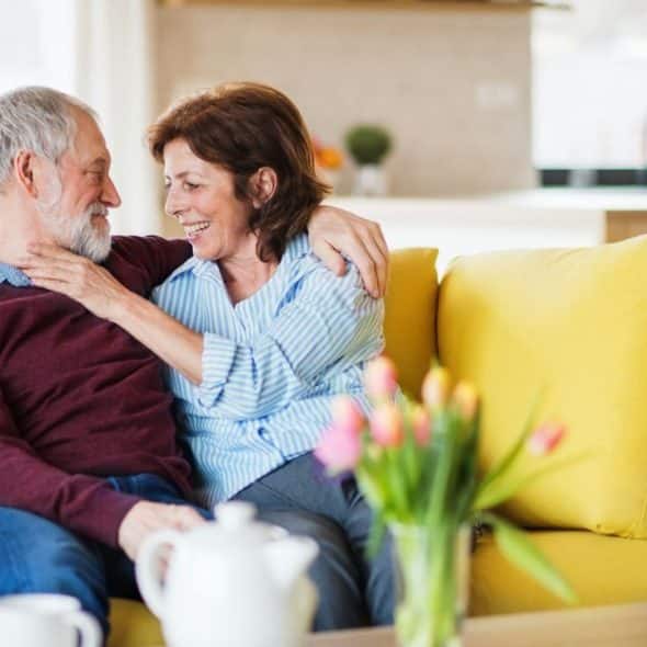 An older couple smiles and hugs on a yellow couch in a brightly lit room.