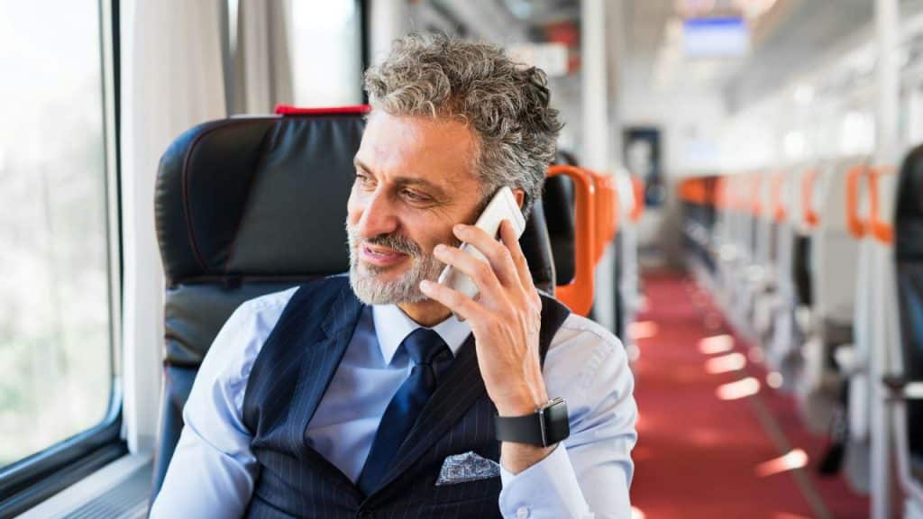 A smiling, gray-haired man in a suit vest is on the phone, looking out a train window.