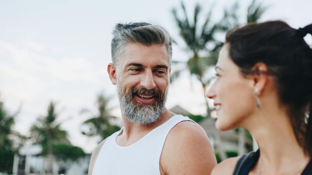 A bearded man in a white tank top smiles at a woman with her hair tied back.