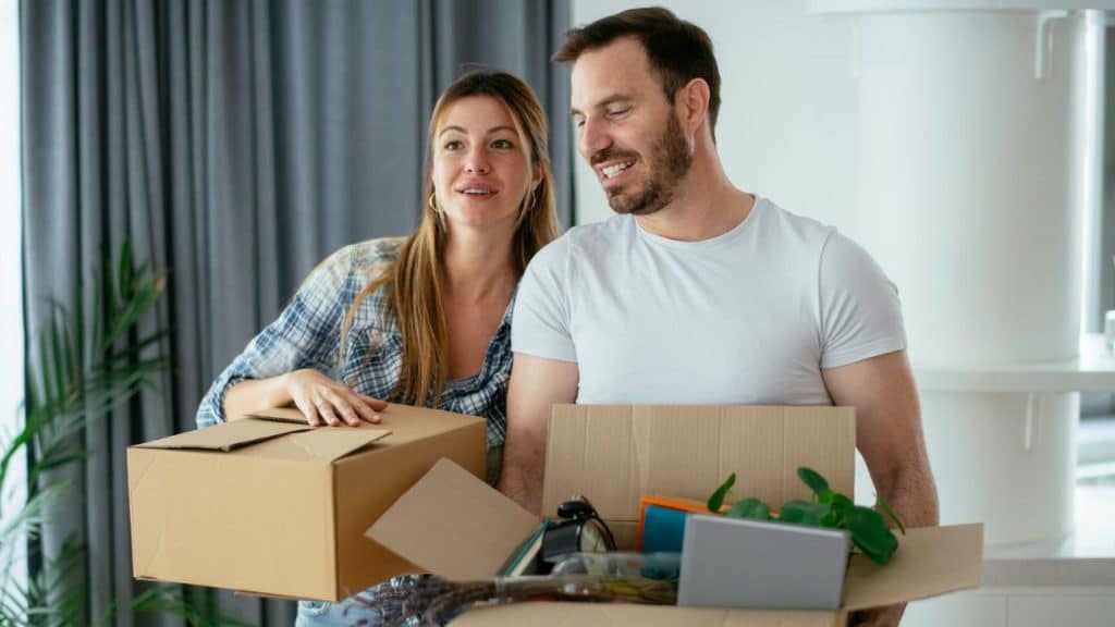 A smiling couple holds moving boxes, looking toward the left.