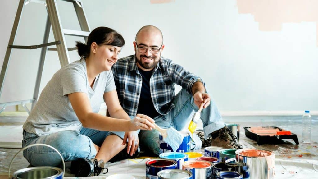 A smiling couple sits on a drop cloth with paint cans and brushes, painting a room.
