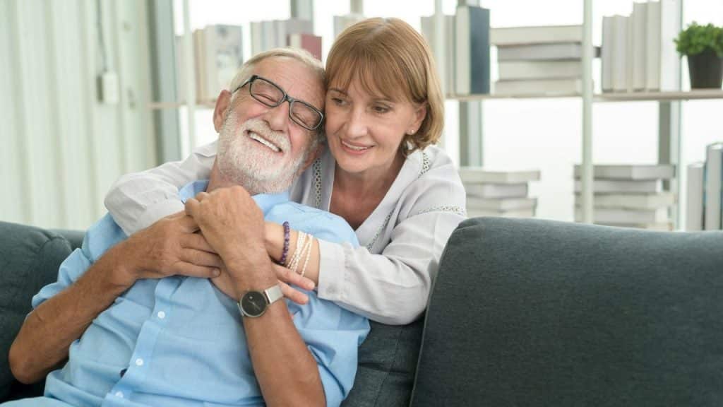 A joyful older couple embraces on a couch, smiling broadly.