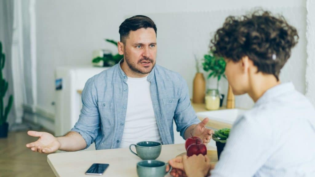 A man with a beard and a light blue shirt talks to a person with curly hair across a table.