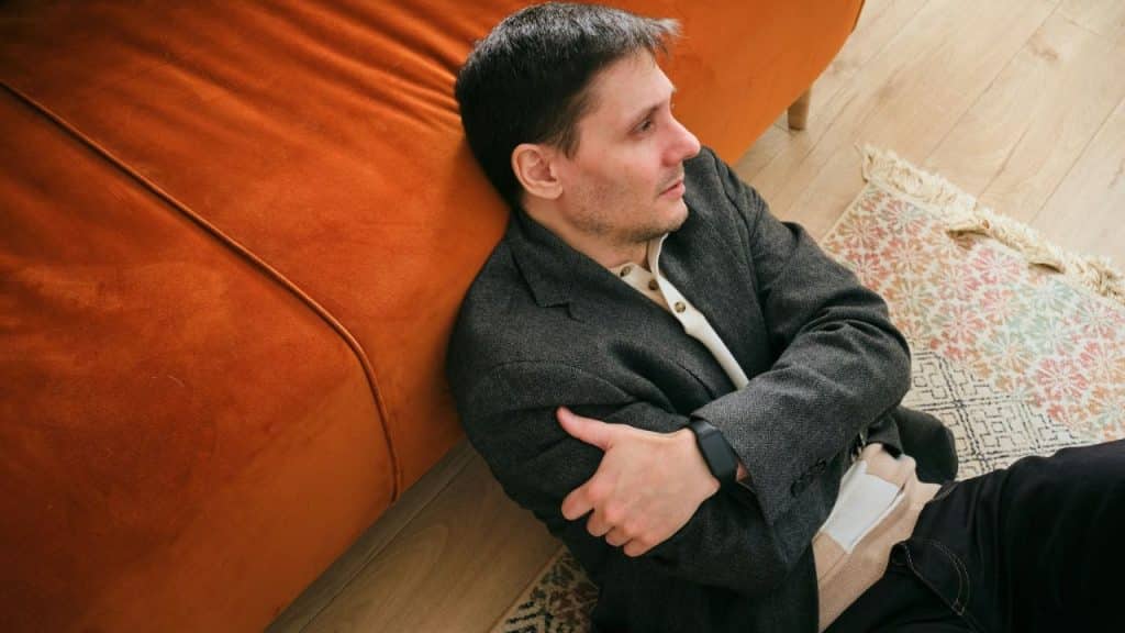 A man in a dark jacket sits on the floor with his arms crossed, looking away from an orange couch.
