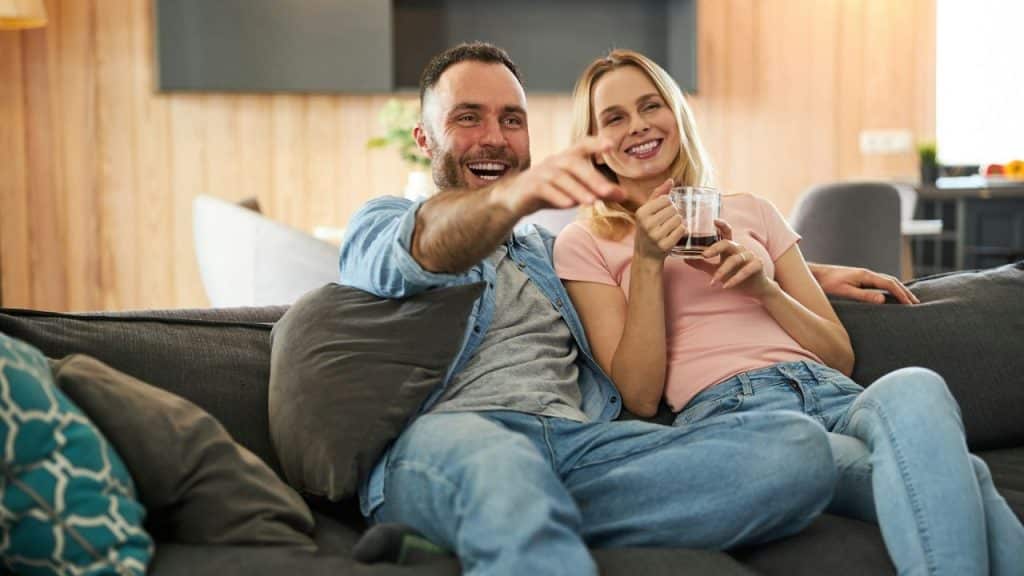 A laughing couple sits on a couch, with the man pointing and the woman holding a cup.