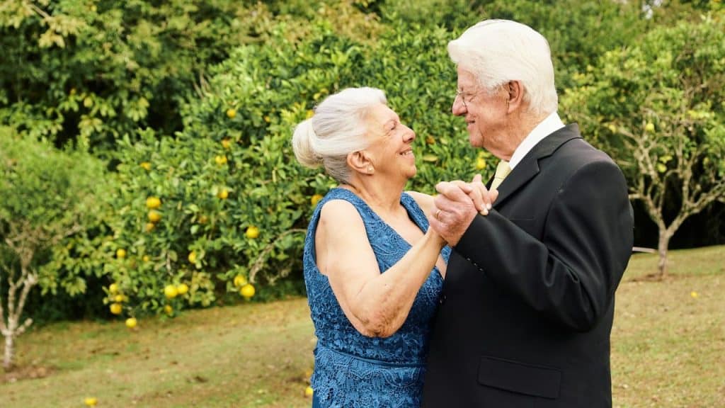 An elderly couple dances outdoors, smiling at each other, with green foliage behind them.