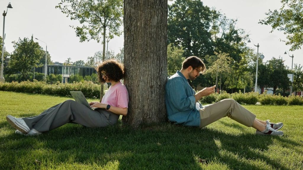 A man and woman lean against a tree, absorbed in their devices, in a sunny park.