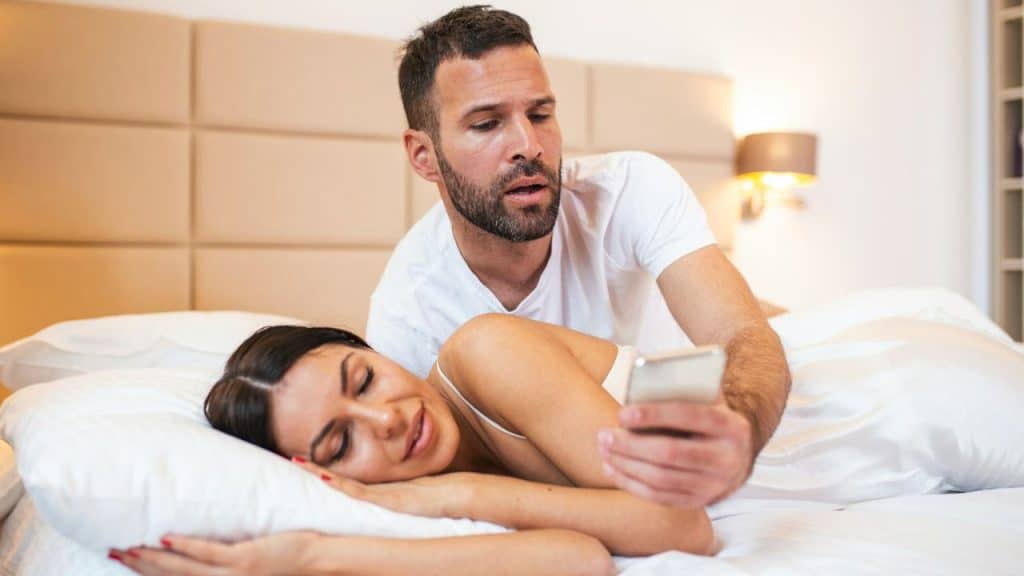A man leans over a woman in bed, looking at her phone.