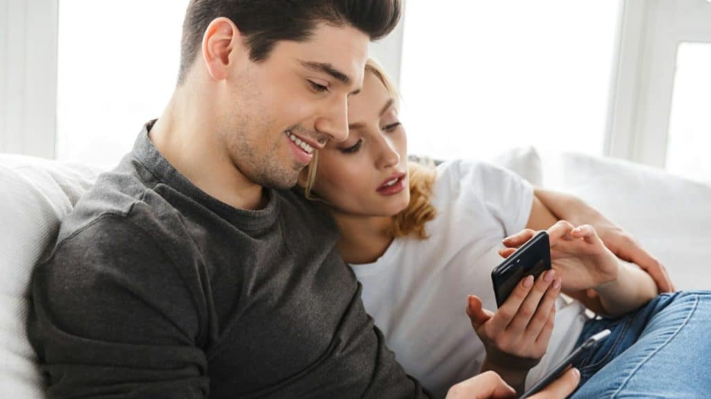A couple on a couch looks at a smartphone, with the woman resting her head on the man.