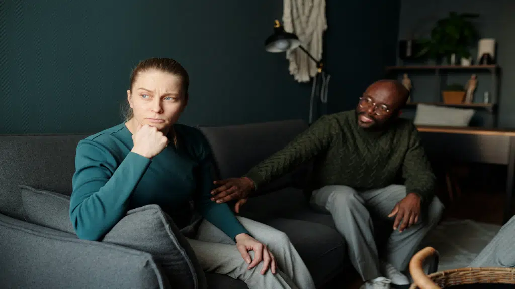 A woman sits on a couch looking upset, while her partner beside her gently reaches out.