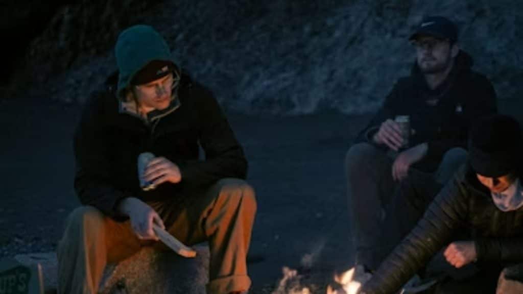 A small group of men sits around a firepit at night, engaged in conversation.