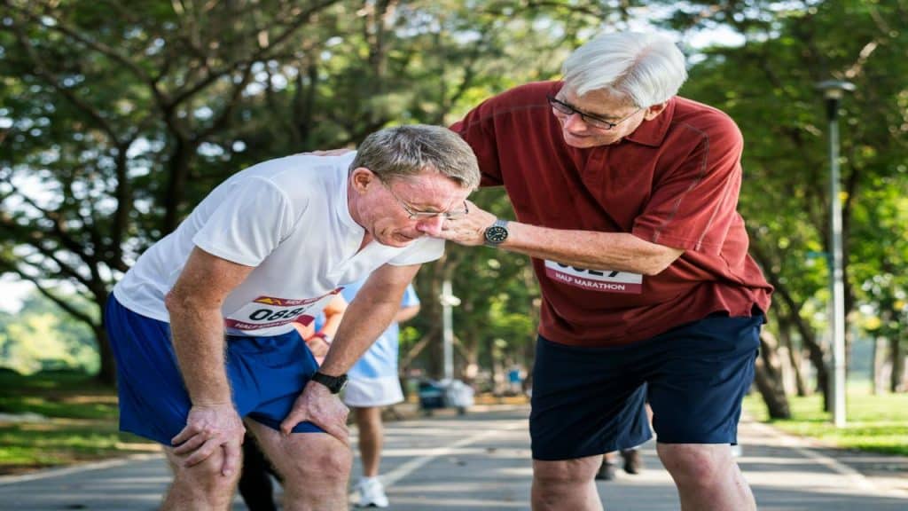A senior man comforting another exhausted runner during a half marathon in a tree-lined park.