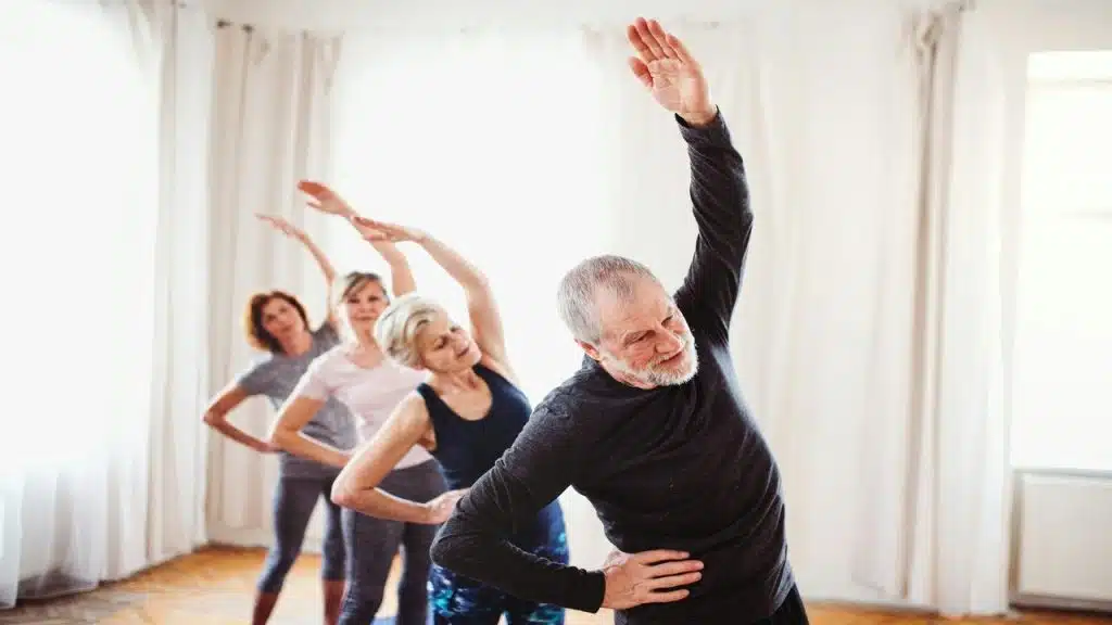 A group of older adults performing standing side stretches in a bright room during a fitness class.