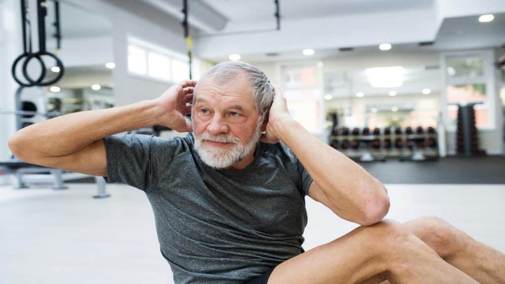 A senior man doing sit-ups on a gym floor with focused determination, surrounded by workout equipment.