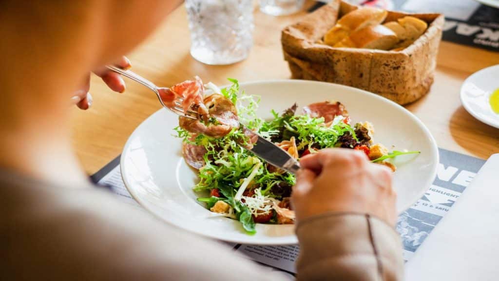 A person eating a fresh salad with cured meats and greens at a restaurant table.
