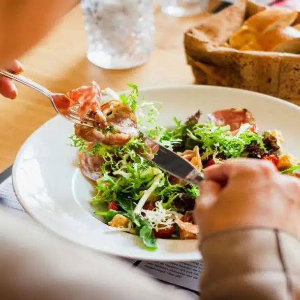 A person eating a fresh salad with cured meats and greens at a restaurant table.
