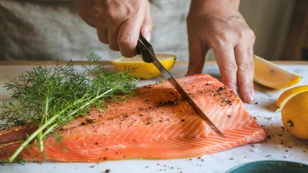 A person slicing a seasoned raw salmon fillet with fresh dill and lemon wedges on a cutting board.
