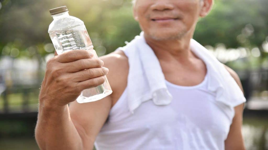An older man in a white tank top holding a bottle of water, with a towel around his neck.