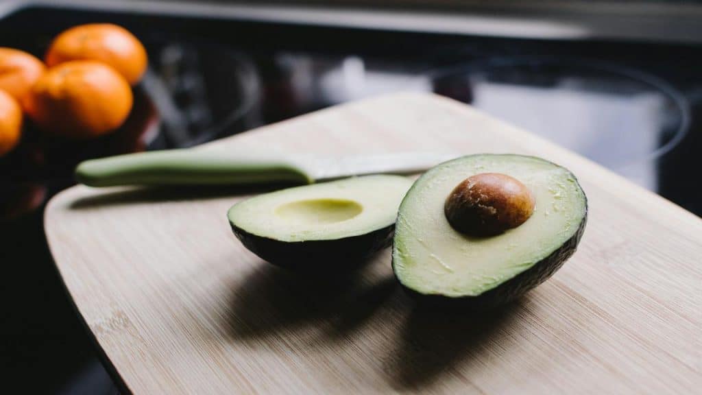 A halved avocado with the pit on a wooden cutting board beside a knife.