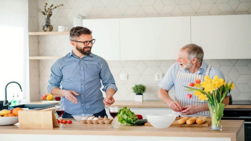 A younger man and an older man smiling while preparing food together in a modern kitchen.