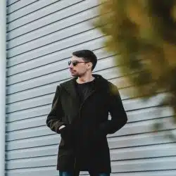 A man in a dark coat and sunglasses standing by a metal wall.