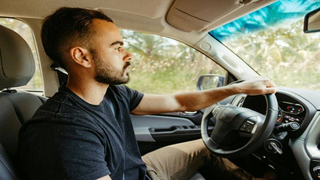 A focused man driving a car through a sunlit, green landscape.