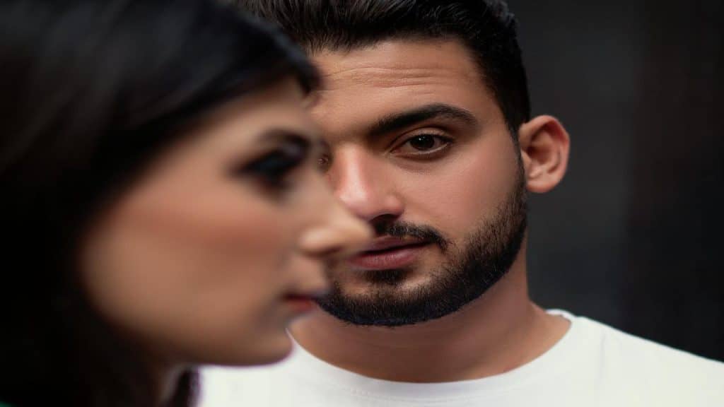 A close-up of a man staring intently while a woman stands in soft focus in the foreground.