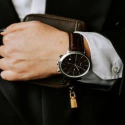 A suited hand wearing a brown leather watch holding a wallet.