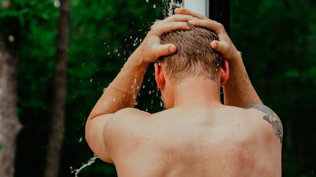 A man from behind takes an outdoor shower, water splashing around his head.