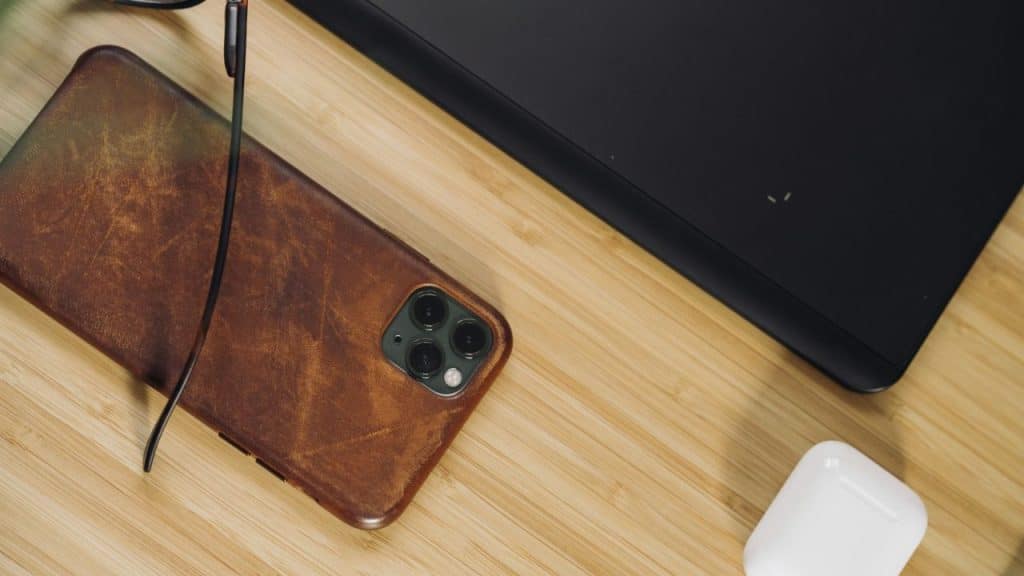 A smartphone in a brown leather case, glasses, and white earbuds are on a wooden desk.
