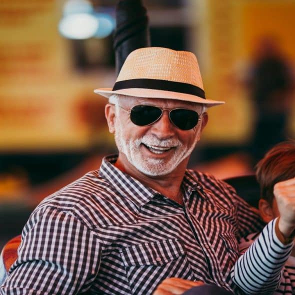 An elderly man in a straw hat and sunglasses smiles joyfully next to a laughing young boy on an amusement park ride.