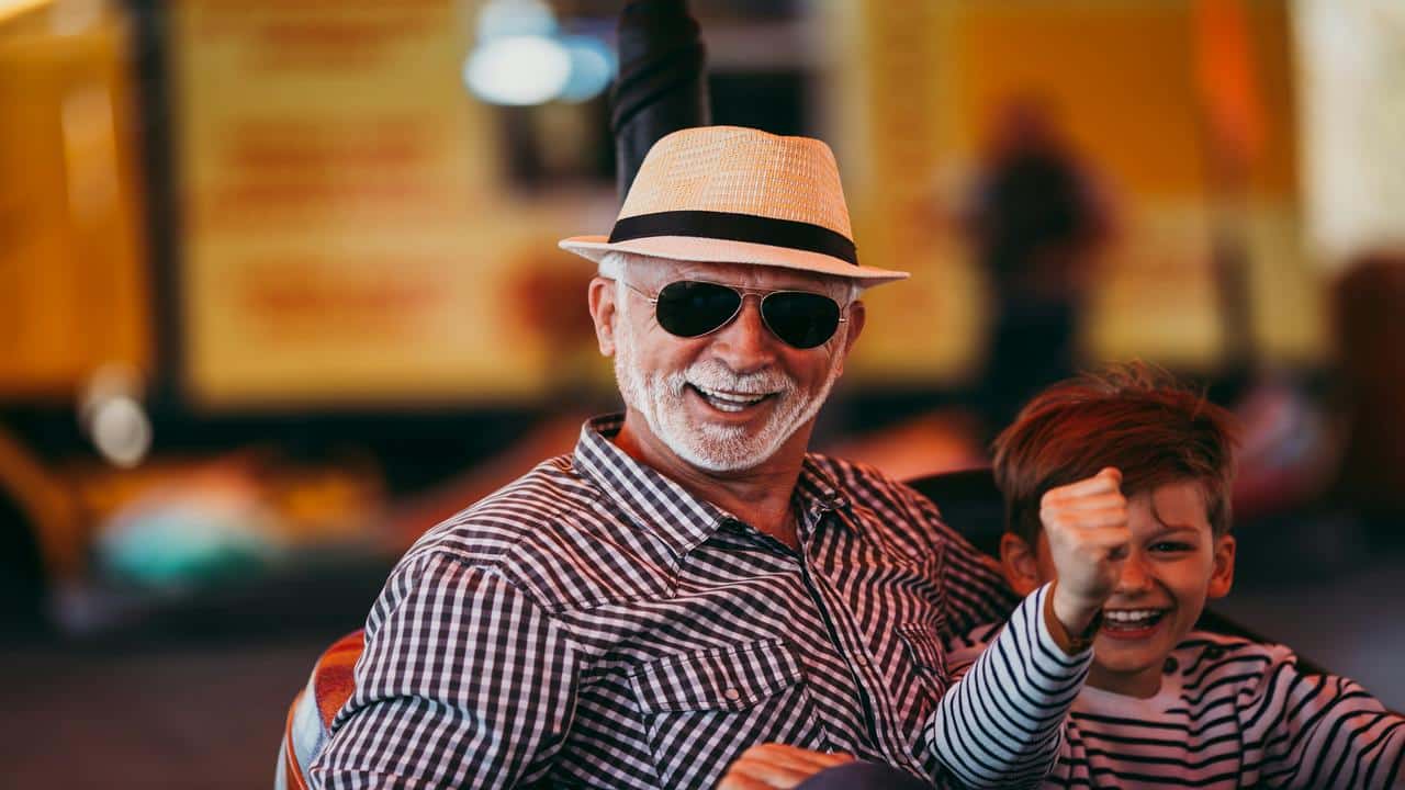 An elderly man in a straw hat and sunglasses smiles joyfully next to a laughing young boy on an amusement park ride.