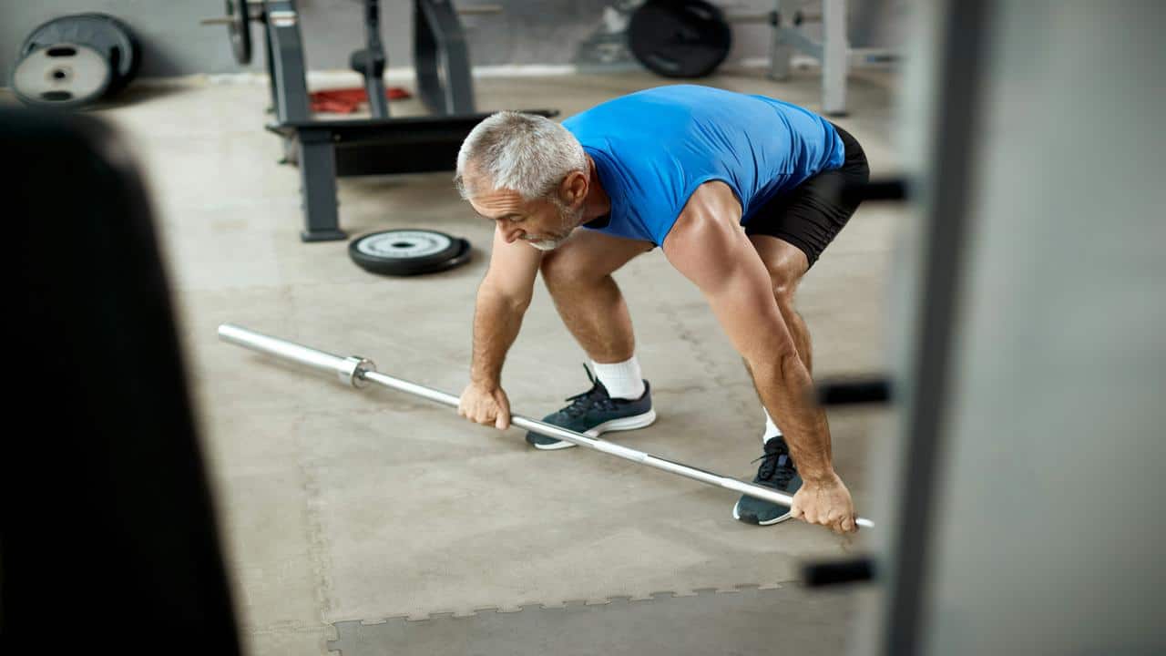A fit older man in a blue sleeveless shirt prepares to lift a barbell in a gym setting.